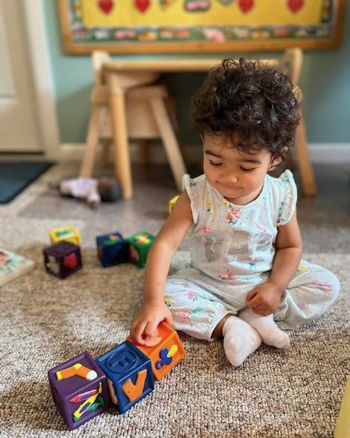 Toddler sitting on carpet, reaching for colorful blocks in a playroom.