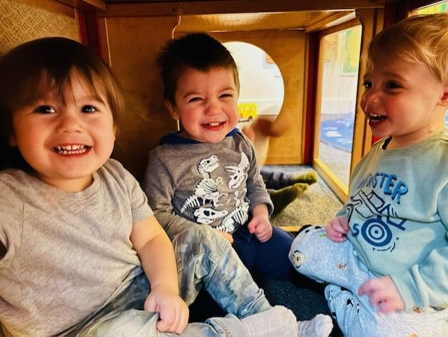 Three smiling children sitting inside a wooden play area, looking at each other.