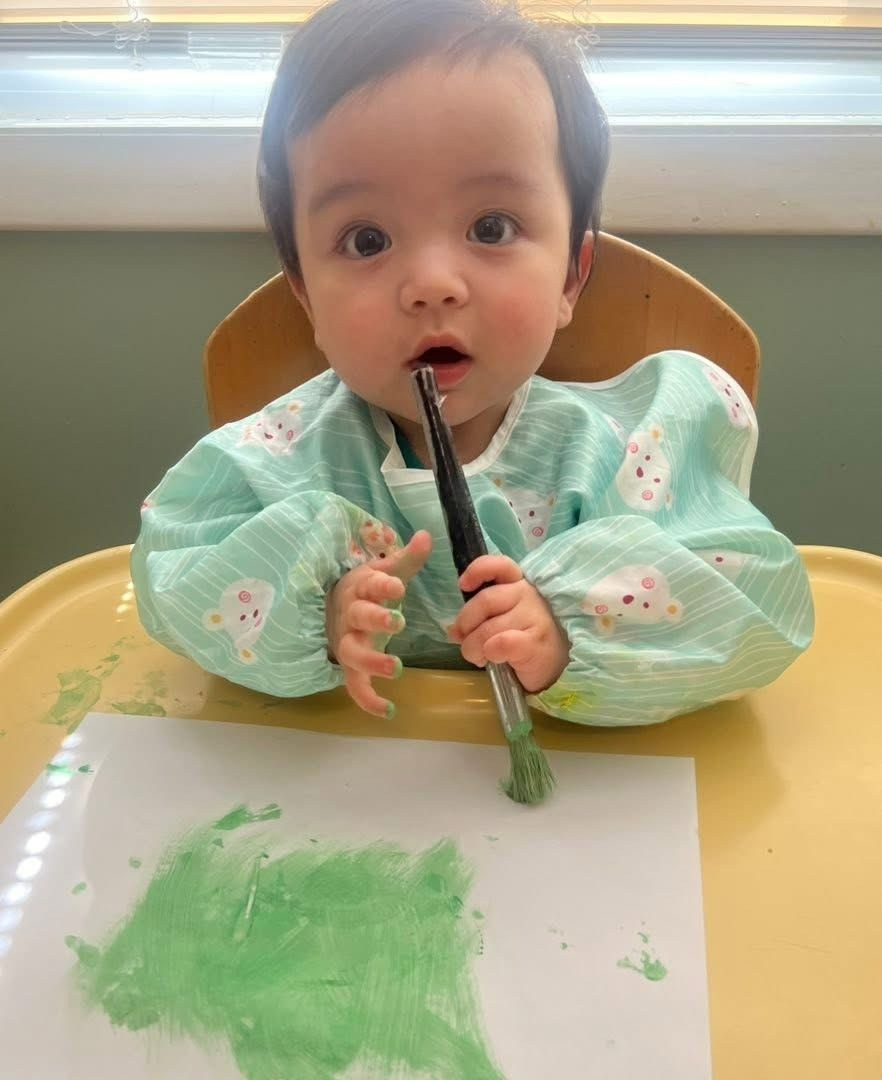 Baby in a mint green outfit painting with a brush at a table, with green paint on paper.