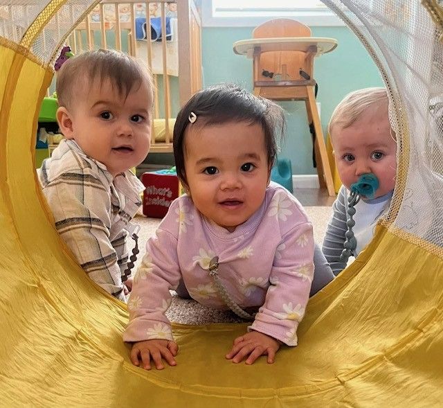 Three babies crawl through a yellow play tunnel, looking toward the camera.