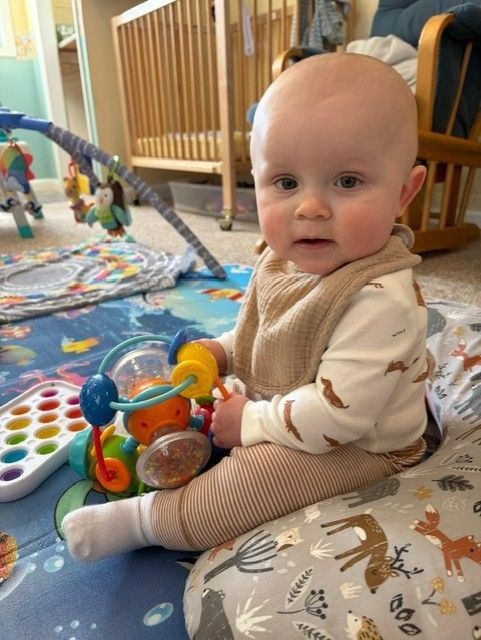 Baby sitting on a play mat with a colorful toy, looking toward the camera.