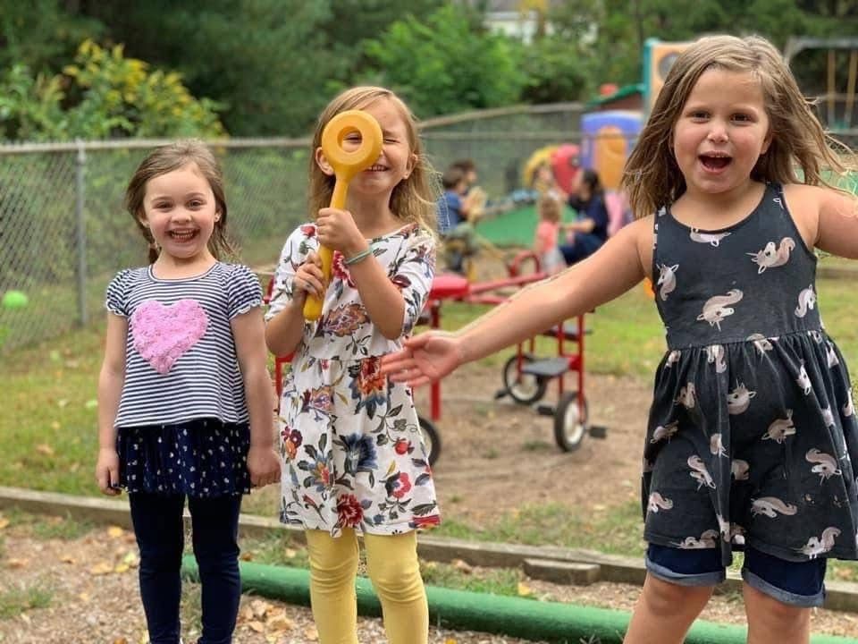 Three children smiling outdoors, one holding a yellow ring, in a park with a stroller behind them