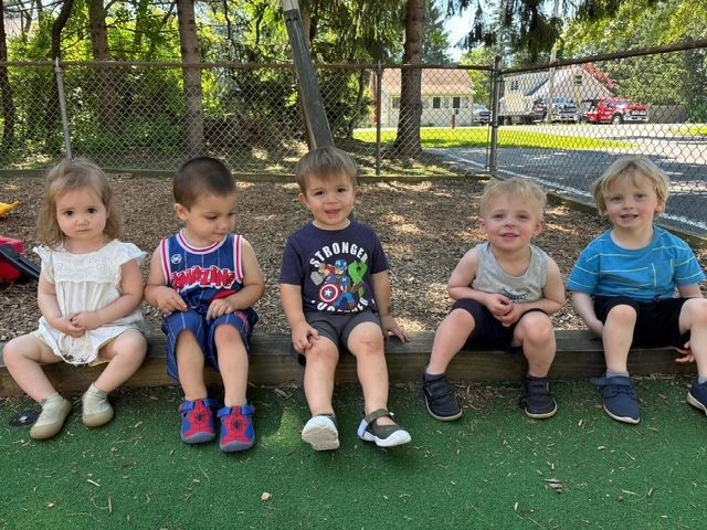 Five young children sitting on a bench outdoors, smiling in a park with a fence and playground behind them