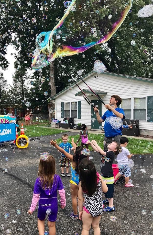 Man blowing a giant rainbow bubble for children in a backyard