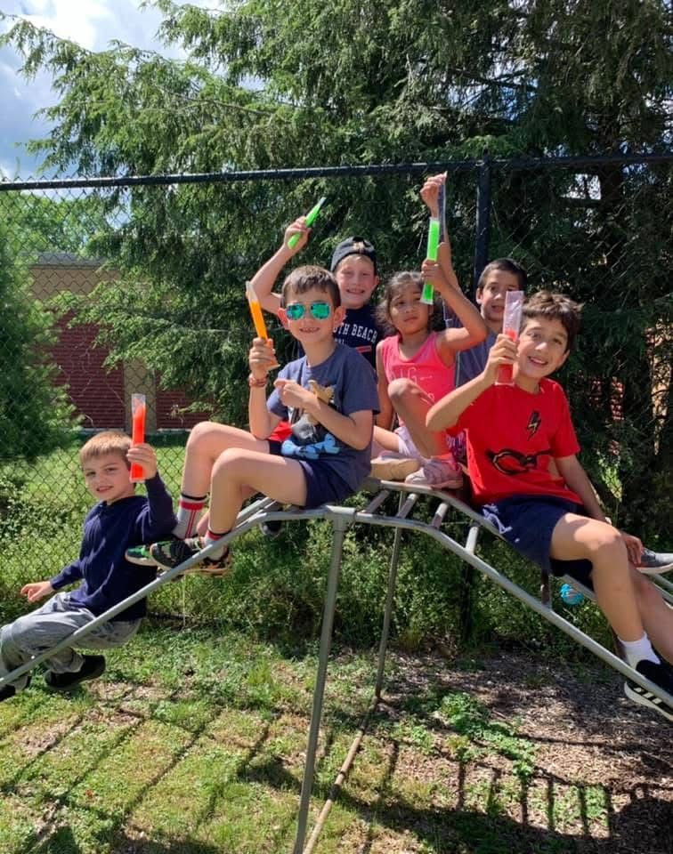 Five children on a metal jungle gym, smiling and waving colorful toy wands in a sunny yard.