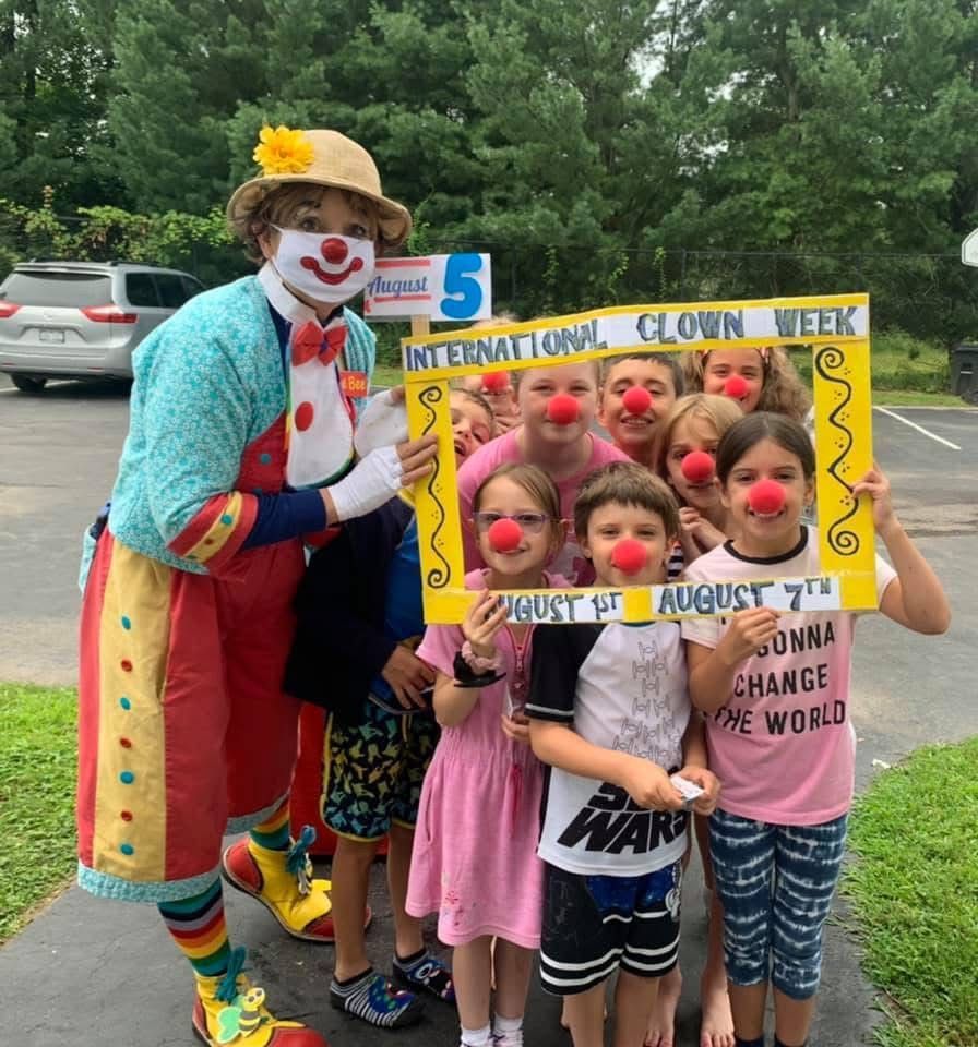 Clown with children holding a “International Clown Week” frame outdoors, smiling with red noses