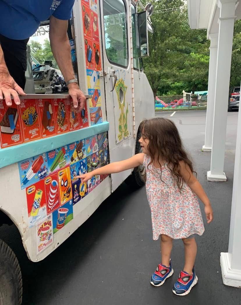 Child in a floral dress reaching toward a colorful ice cream truck at a curbside stop