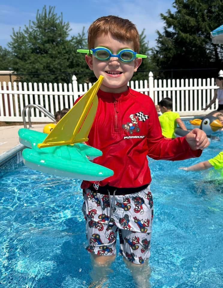 Child in swim goggles holding a toy shark in a pool, wearing a red jacket and patterned swim shorts.