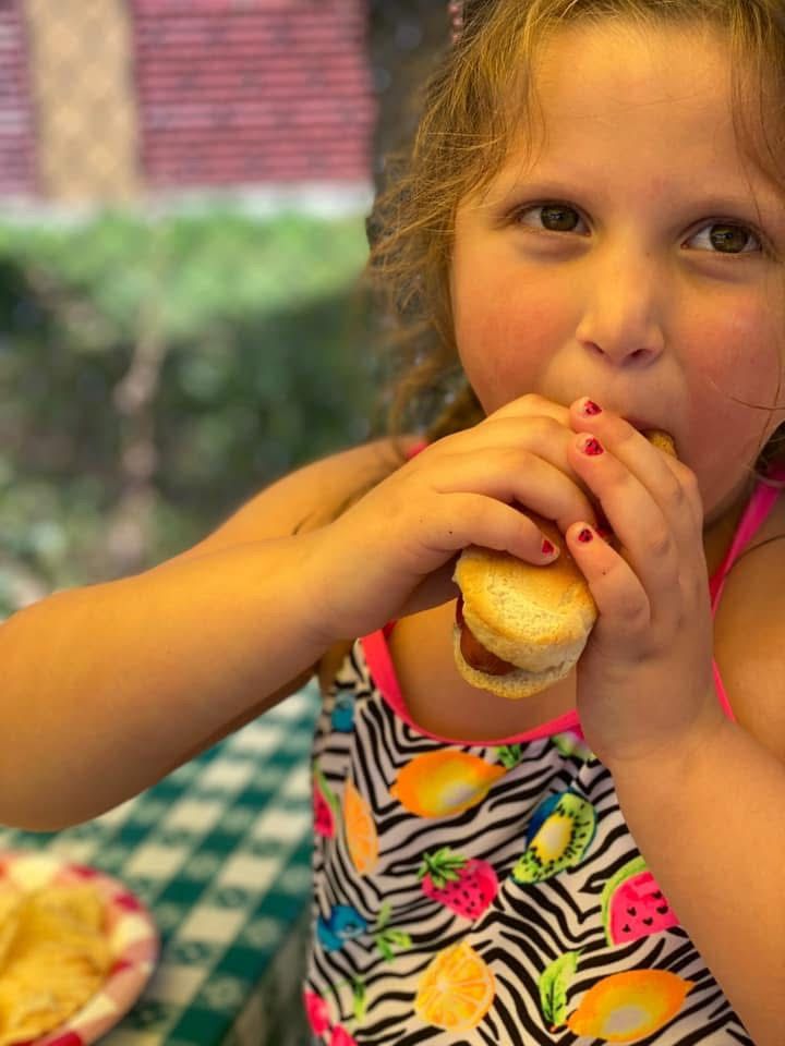 Child eating a hot dog at a table, with a zebra-print top and blurred greenery in the background