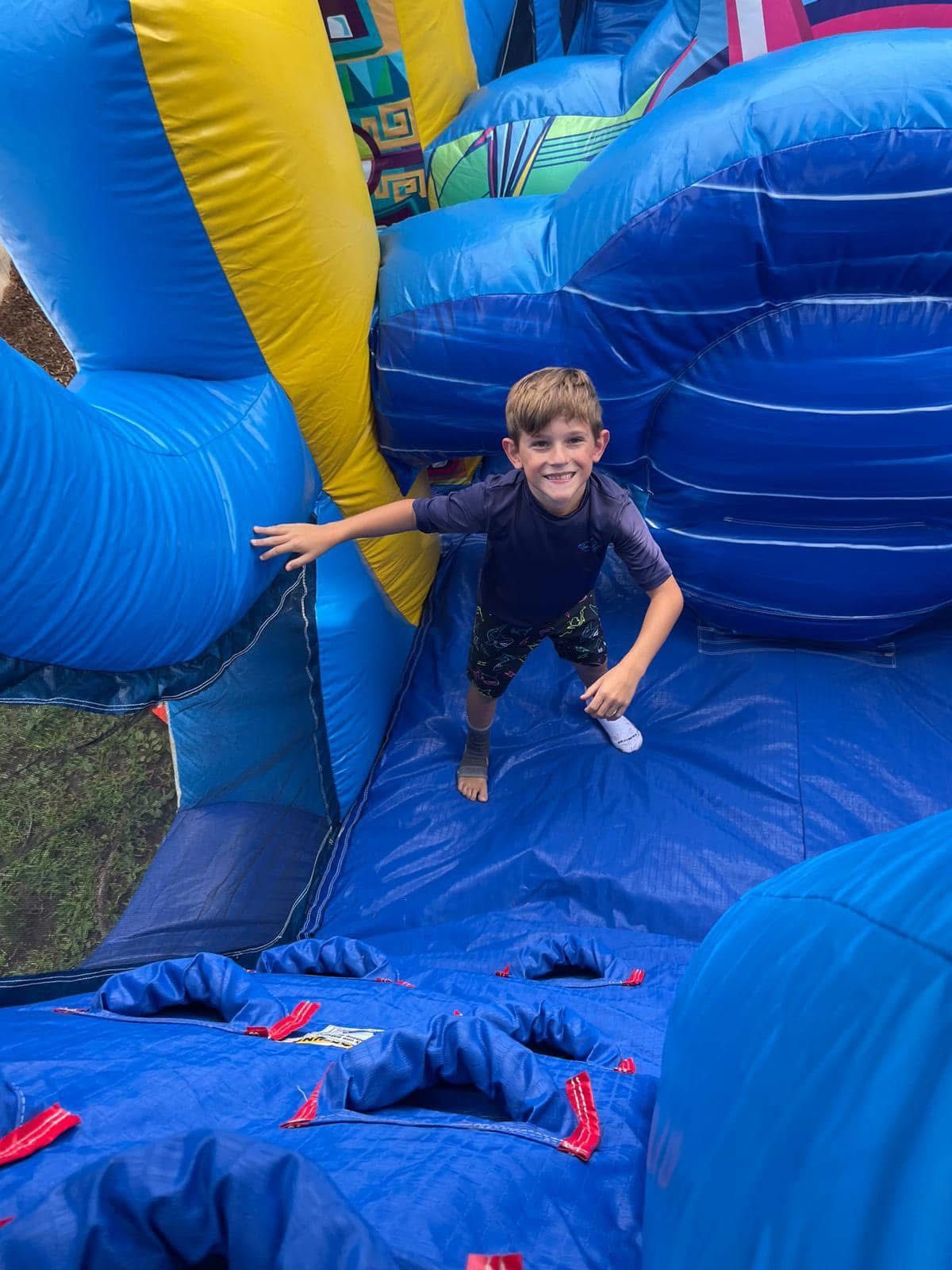 Child climbing a blue-and-yellow inflatable obstacle course, smiling with one hand on the side wall.