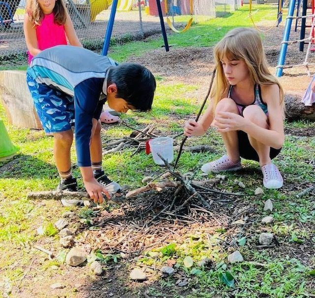 Children tending a small fire pit outdoors with sticks in a grassy playground area