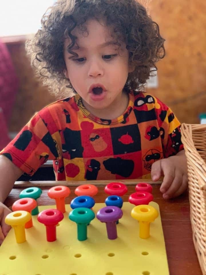 Child playing with colorful peg toy on a table, mouth open in concentration.
