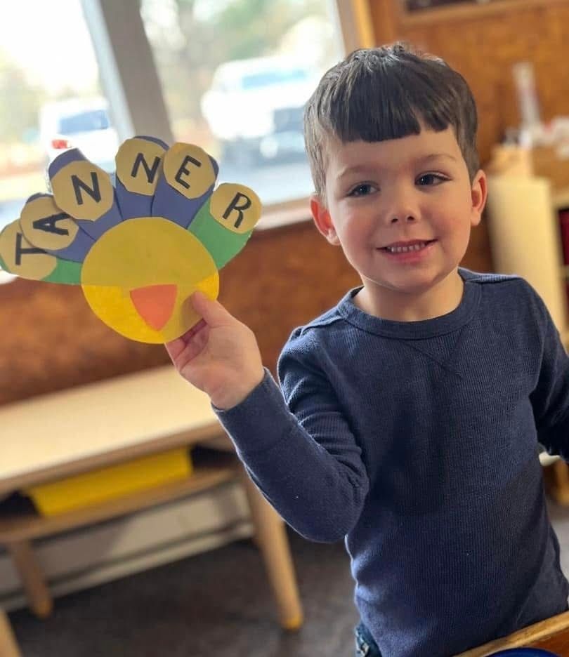 Child holding a colorful flower craft with the name “TANNER” in a classroom setting