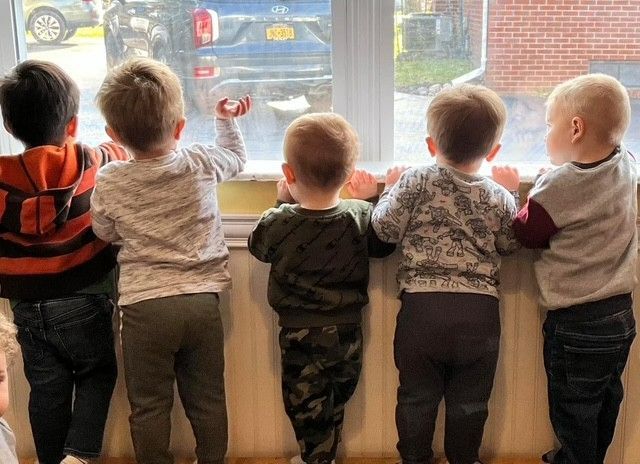 Five children stand on a windowsill, looking outside at a rainy street.