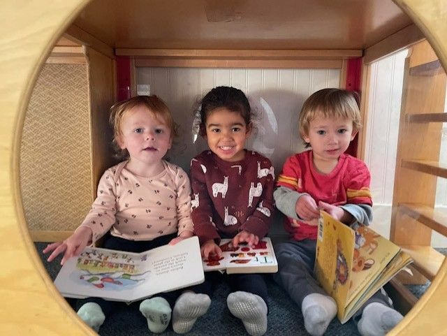 Three children sitting in a cozy wooden playhouse, holding books and looking at the camera.