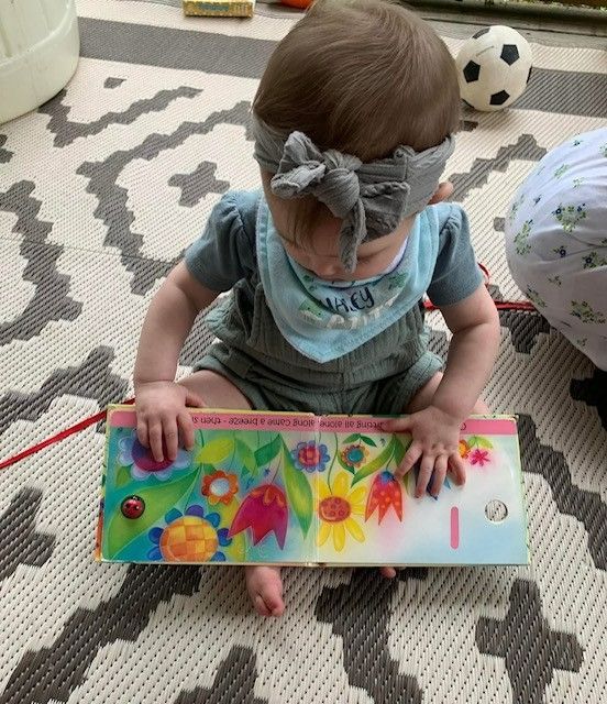 Toddler painting a colorful board on a patterned rug, wearing a gray bow headband.