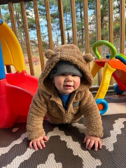 Toddler in a brown bear costume crawling on a play mat at an outdoor playground.