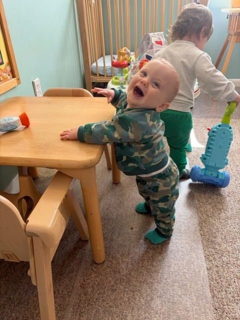Toddler standing at a small table in a playroom, mouth open, with another child nearby.