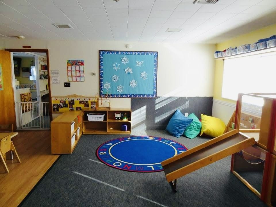 Childcare playroom with a blue rug, toy shelves, cushions, and a slide by the window