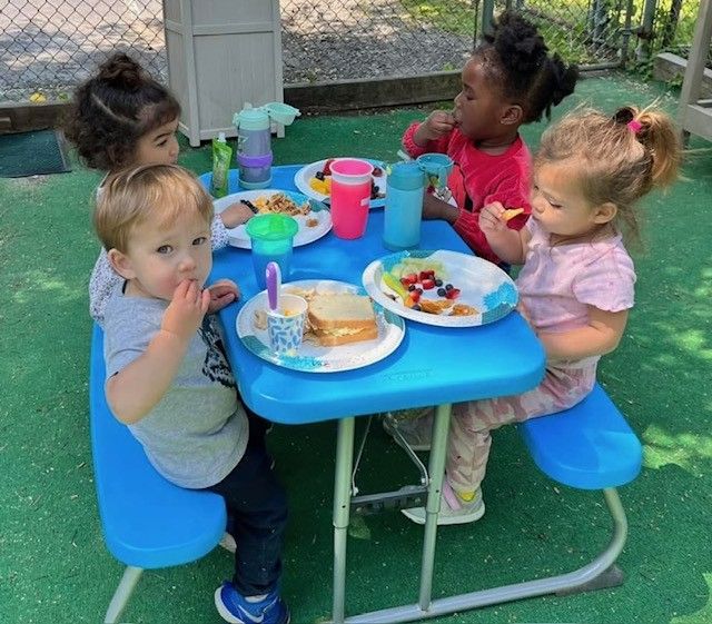 Four toddlers sit at a blue picnic table, eating snacks and drinks outdoors.