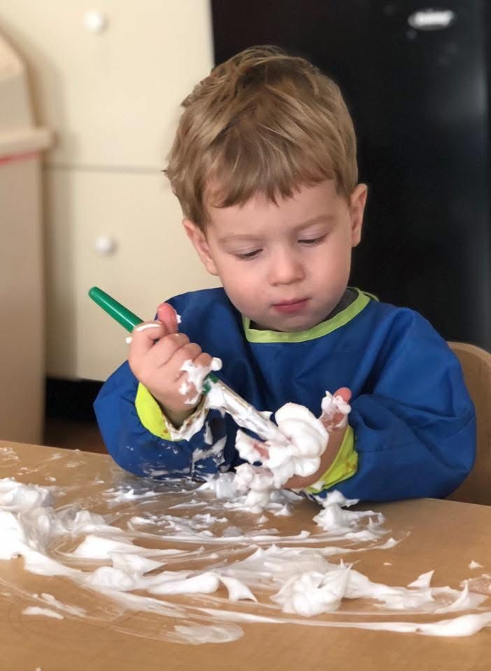 Child at a table, holding a green marker and covered in white cream, concentrating while playing with it.