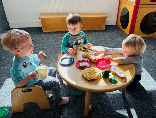 Three toddlers sit around a round table with colorful dishes in a playroom.