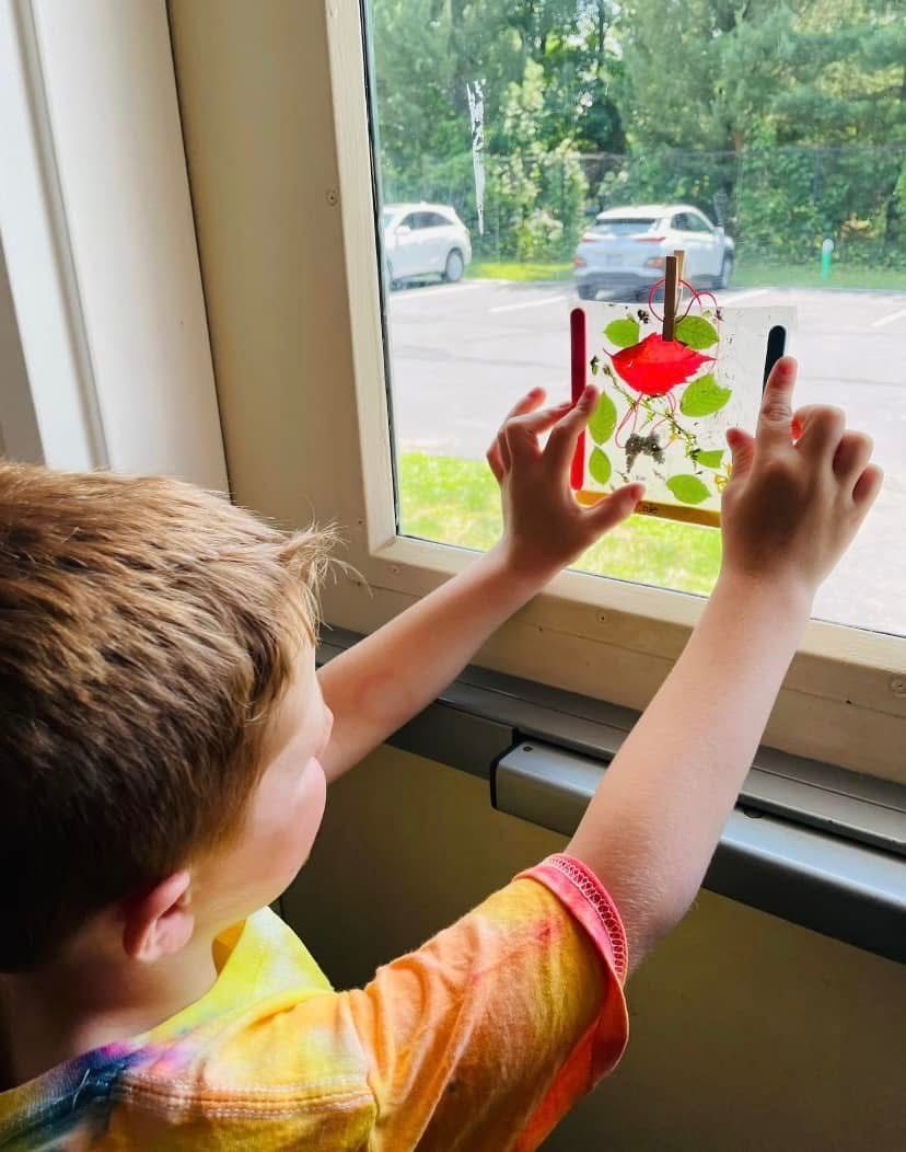 Child reaching toward a window with a colorful heart decoration and two candles, looking outside.