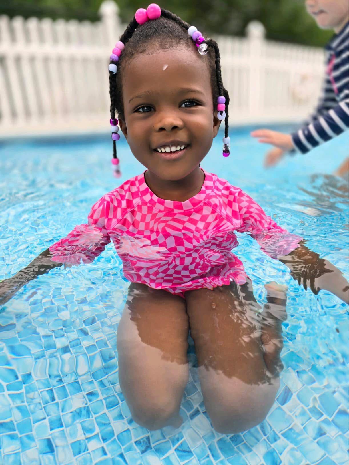 Child smiling in a pink swimsuit kneeling in a pool with blue water, holding the edge