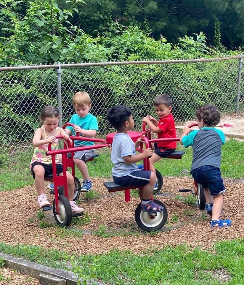 Children riding a small merry-go-round in a grassy playground beside a chain-link fence.