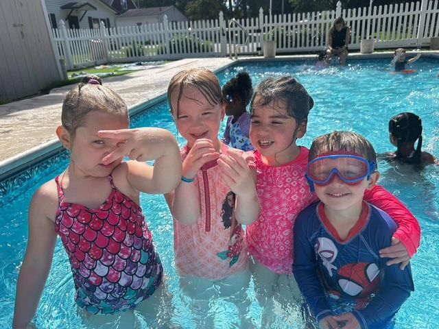 Four children in colorful swimsuits pose and splash in a backyard swimming pool.