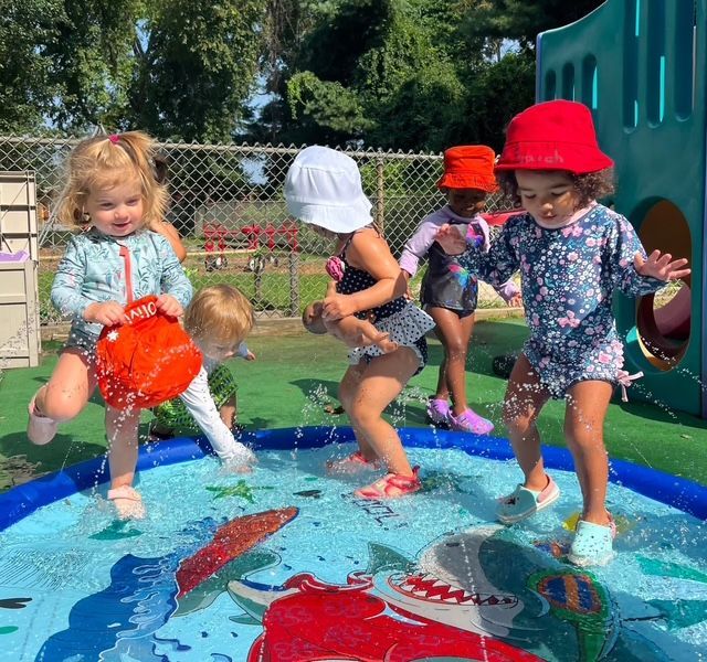 Children splashing in a shallow outdoor water play area with colorful hats and toys