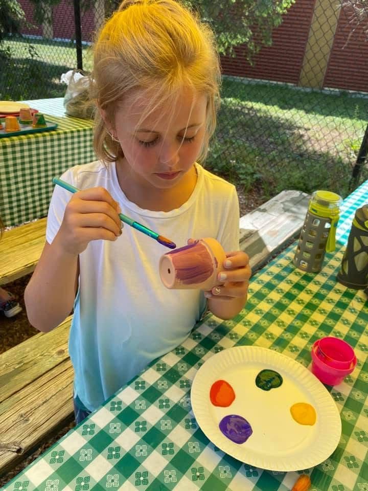 Child painting a small pot at an outdoor table with colorful paint on a paper plate