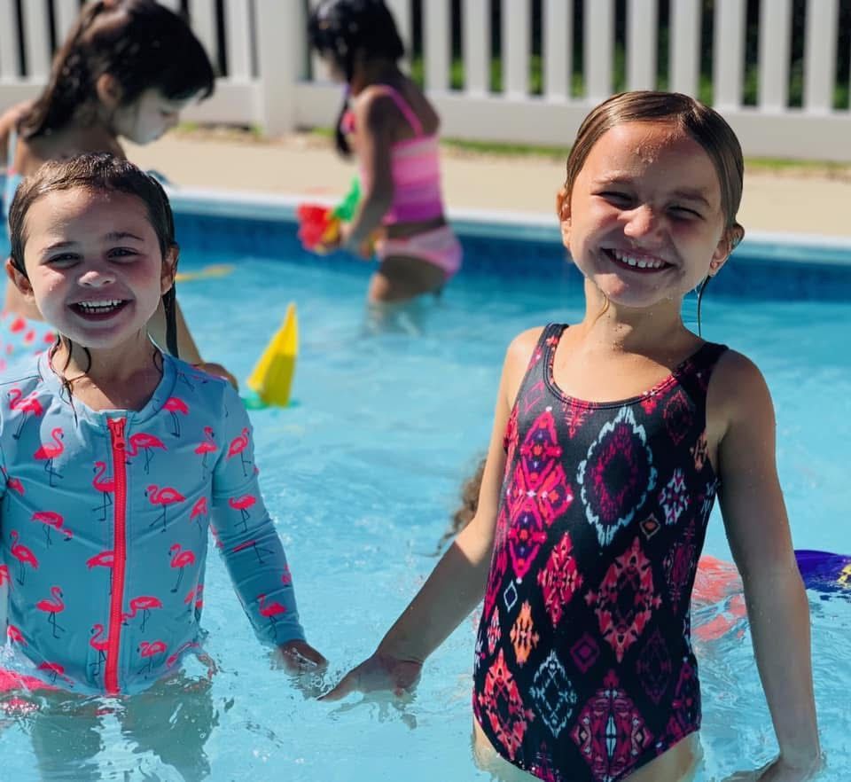 Two children smiling and holding hands in a swimming pool, wearing colorful swimsuits.