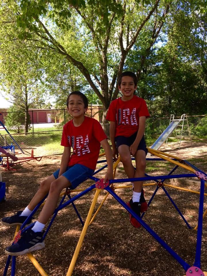 Two children in red shirts sit on a blue-and-yellow jungle gym in a park.