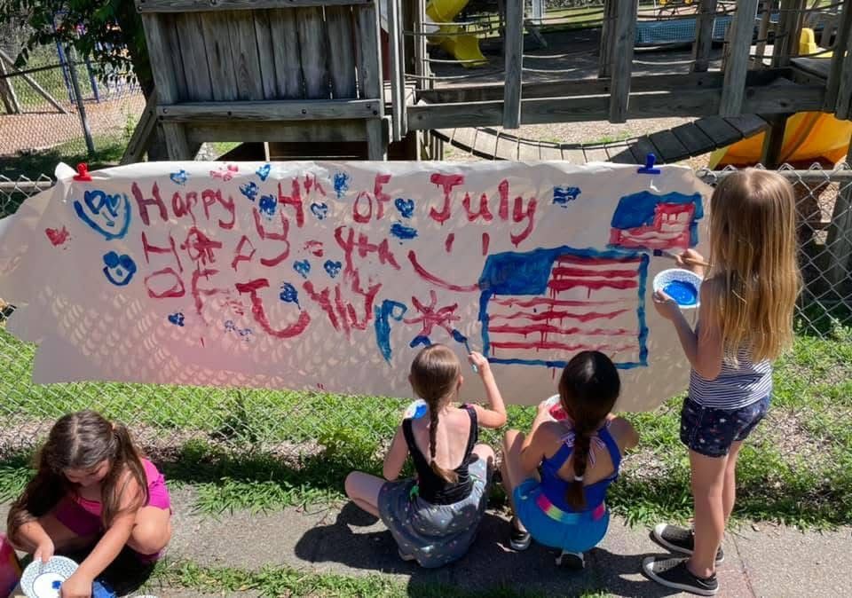 Children painting a long outdoor banner with red and blue July 4th decorations