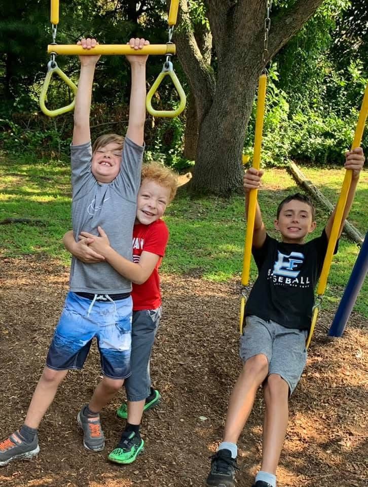 Three children laughing on playground swings under a tree, one hanging from a bar and two on yellow swings.
