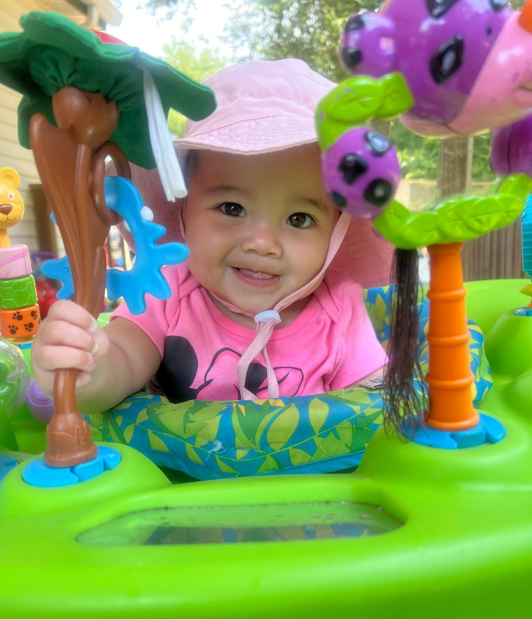 Smiling baby in a pink shirt sitting in a colorful activity walker with toy animals and trees