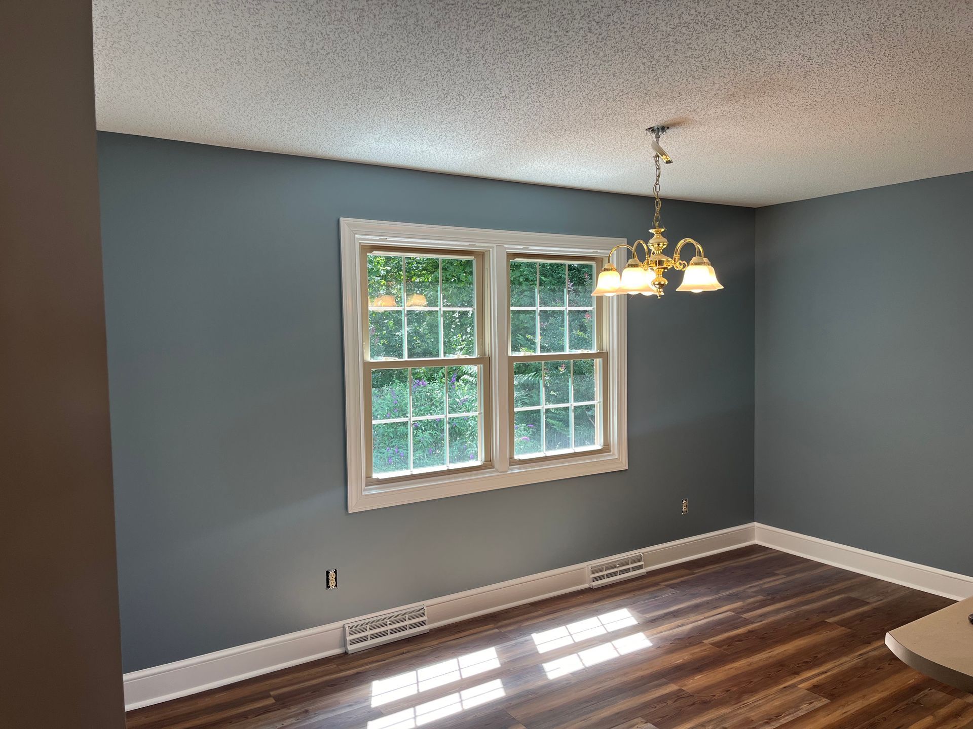 Room with blue walls, a window overlooking greenery, wood floor, and chandelier.