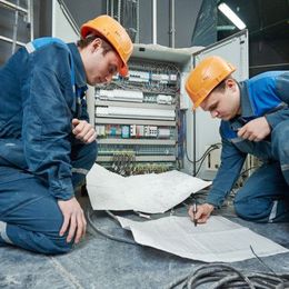 Two electricians in blue coveralls and orange hard hats review wiring diagrams beside an open control panel.