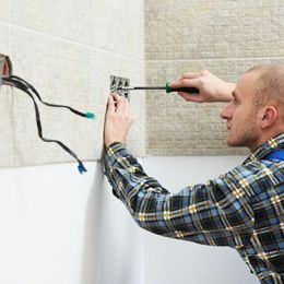 Man wiring an electrical outlet with a screwdriver beside exposed wires on a tiled wall