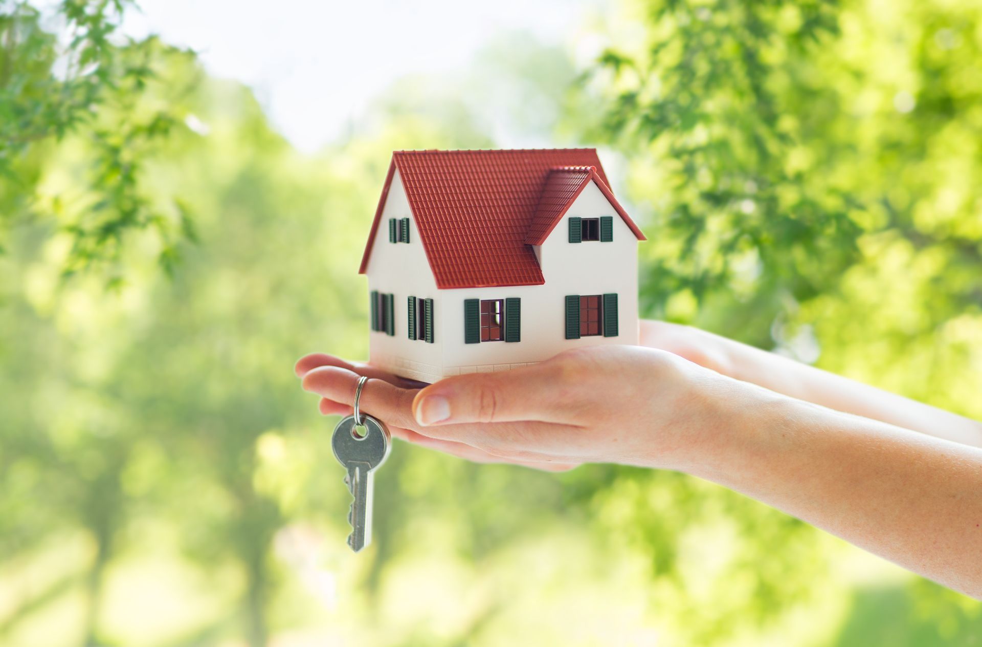 Mani che tengono una casa in miniatura con il tetto rosso e la chiave, su uno sfondo verde sfocato.