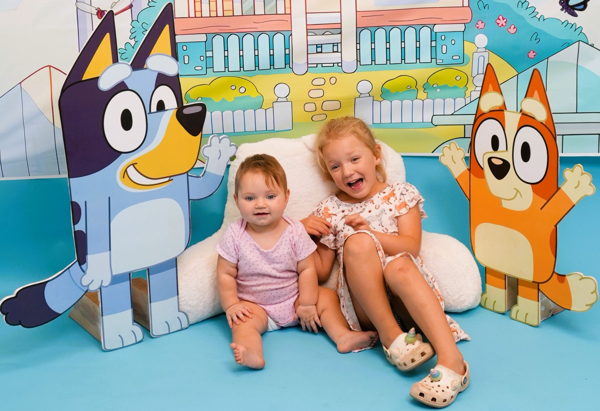 Two young children seated in front of Bluey and Bingo cutouts, smiling. Light blue background.