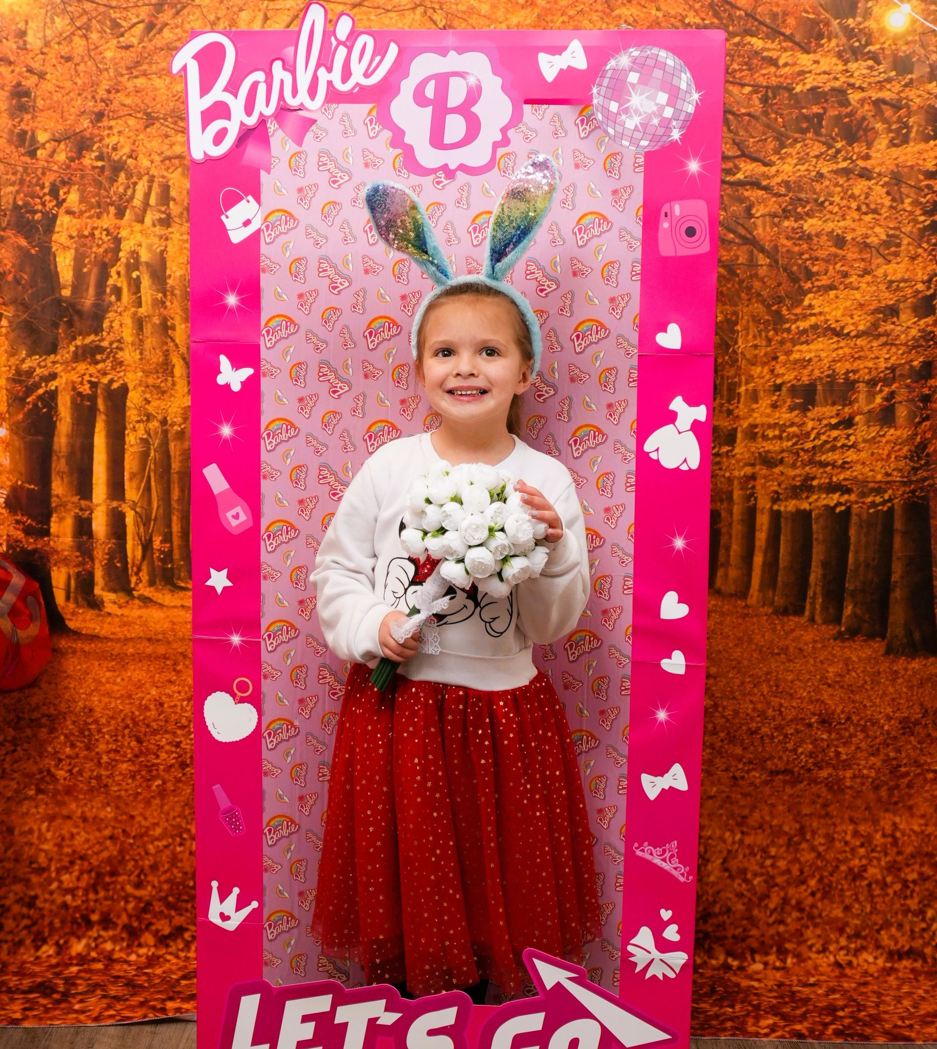 Girl posing in a Barbie-themed photo frame, wearing bunny ears and a red skirt, with an autumn forest backdrop.