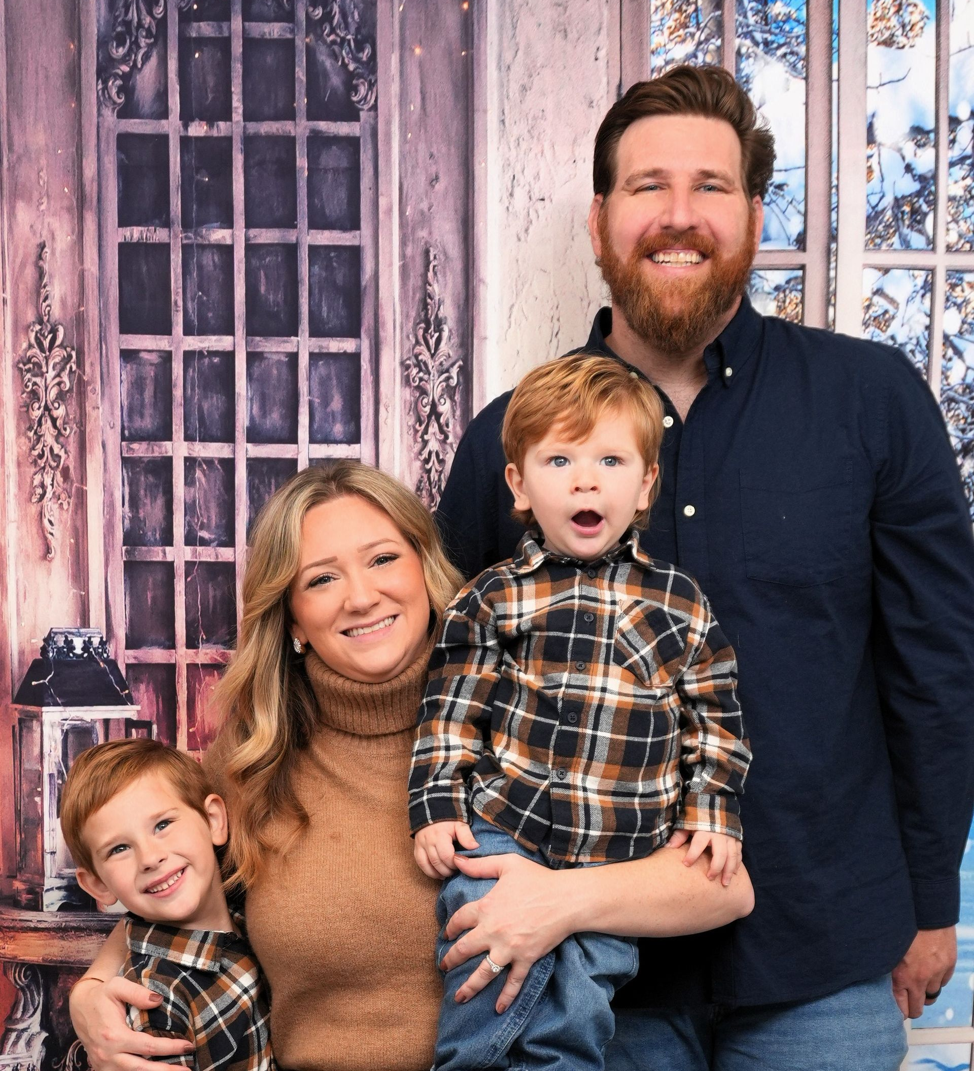 Family of four poses in front of a winter-themed backdrop; the two boys wear matching plaid shirts, and the parents smile.