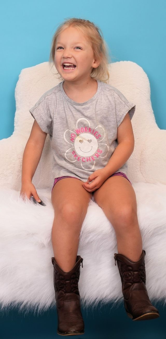 Child seated on a fluffy white surface, wearing a grey shirt, brown boots, and smiling broadly. Turquoise background.