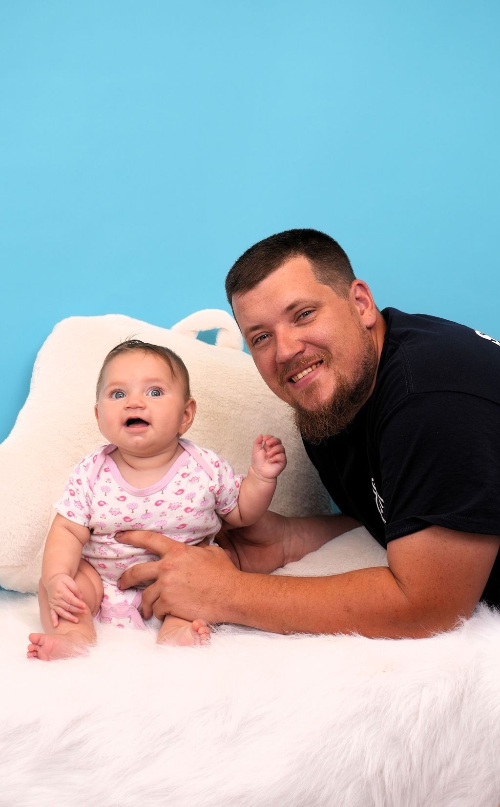 Father and baby sit together on a furry white surface against a blue backdrop. The baby smiles.