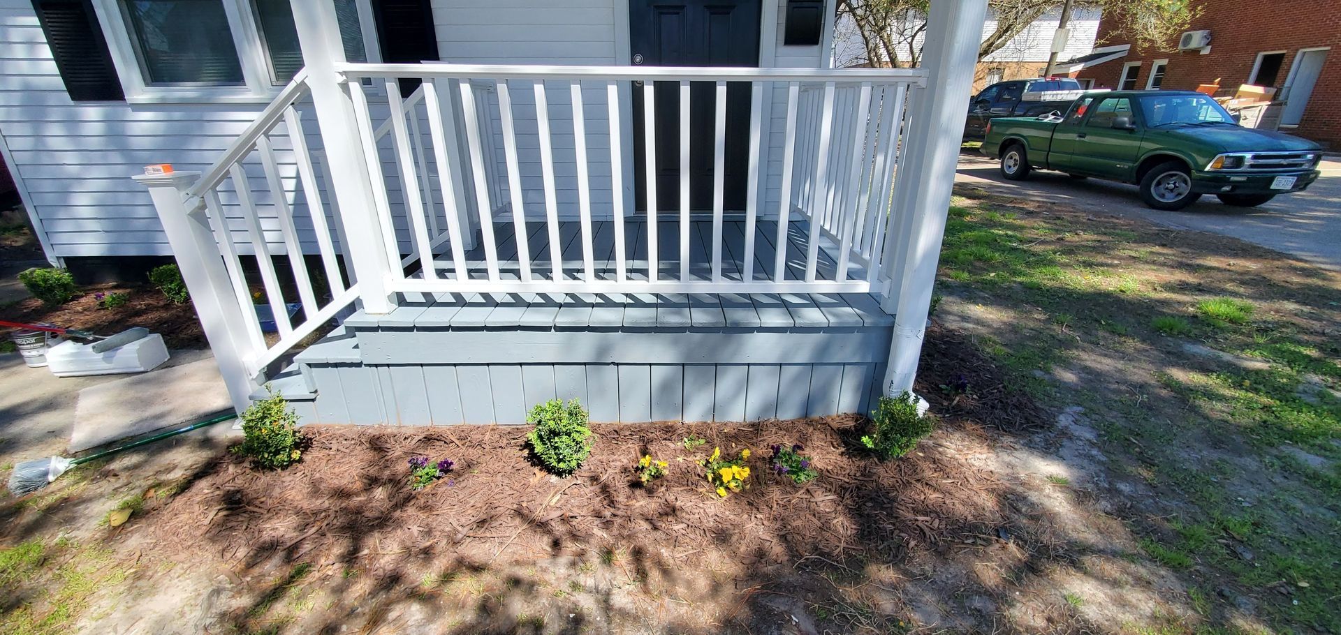 Front porch with white railing, gray siding, and small plants in a mulched garden bed.
