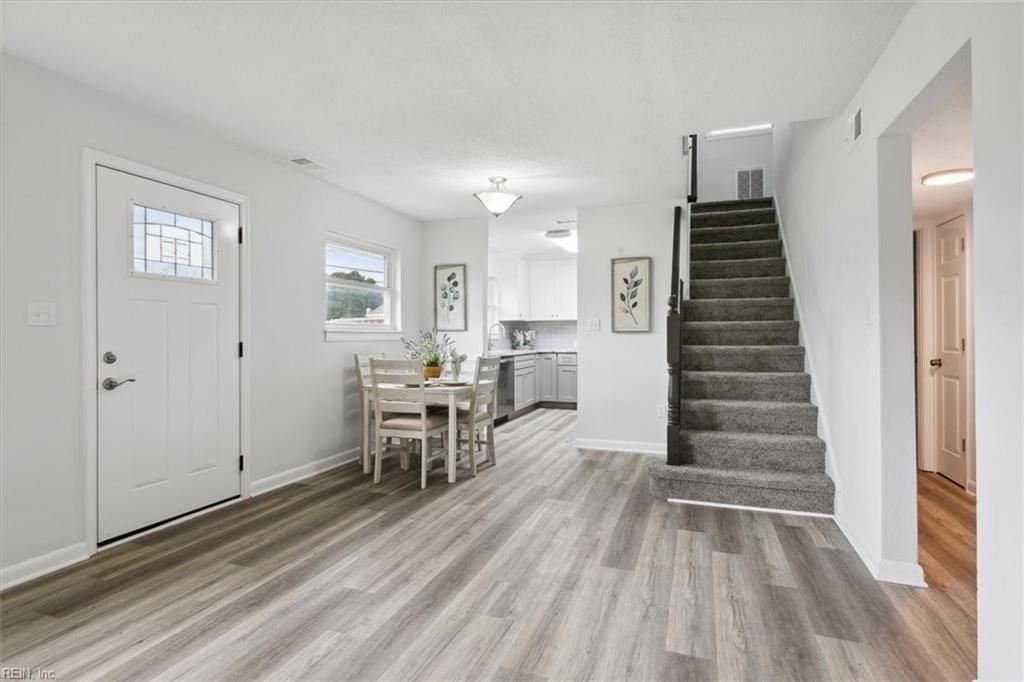 Bright open dining area with white walls, gray wood floors, staircase, and kitchen in the background