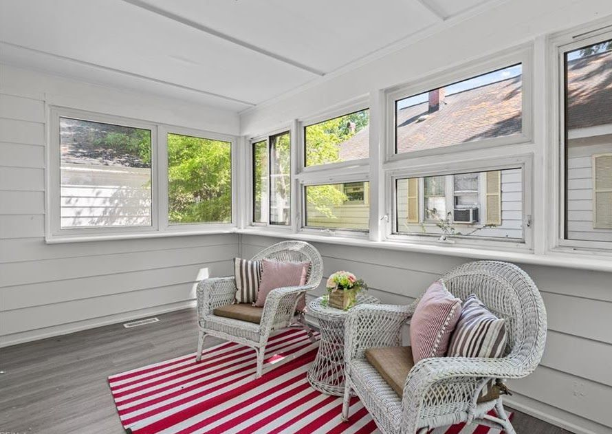Bright sunroom with white wicker chairs, pink pillows, and a red-striped rug by large windows.