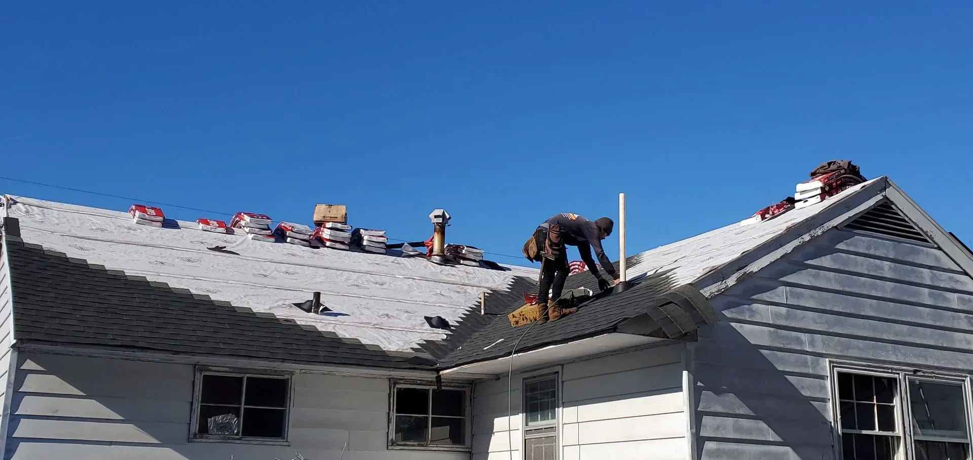 A person is on a roof of a house, possibly repairing or replacing shingles under a clear blue sky.